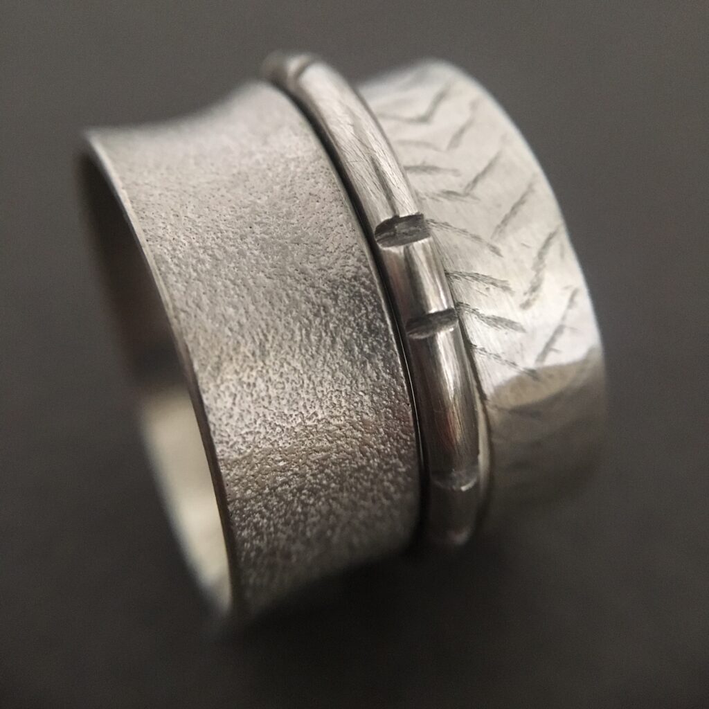 A close-up of two textured silver stacking rings, one featuring a wide matte finish and the other a narrower band with an engraved chevron pattern.